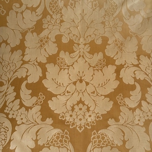 Puede incluir: Primer plano de una tela dorada con un patrón de damasco floral repetido. El diseño presenta grandes flores y hojas estilizadas en un tono dorado más claro, creando un aspecto texturizado y elegante. Ideal para tapicería o cortinas.