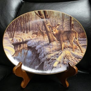 May include: An oval decorative plate featuring a winter forest scene with deer. The plate has a gold rim and is displayed on a wooden stand. The artwork depicts a snowy landscape with a stream and several deer.
