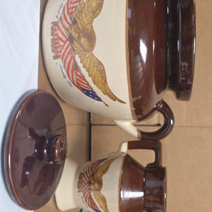 May include: A brown and cream ceramic pitcher and creamer set featuring an American flag and eagle design. The pitcher has a handle and a lid. The creamer has a handle and a spout. The text "Seventy Six" is visible.