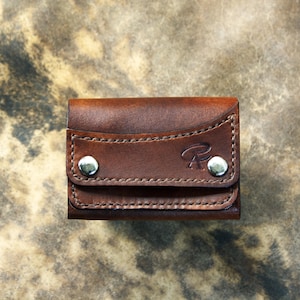 Può includere: Un piccolo portafoglio in pelle marrone con una patta fissata da due bottoni a pressione argentati. Il portafoglio presenta cuciture decorative e un sottile design in rilievo. Il design generale è compatto e sembra fatto a mano.