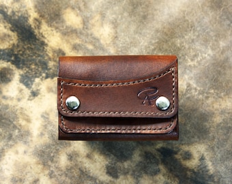 May include: A small, brown leather wallet with a flap secured by two silver snaps. The wallet features decorative stitching and a subtle embossed design. The overall design is compact and appears to be handmade.