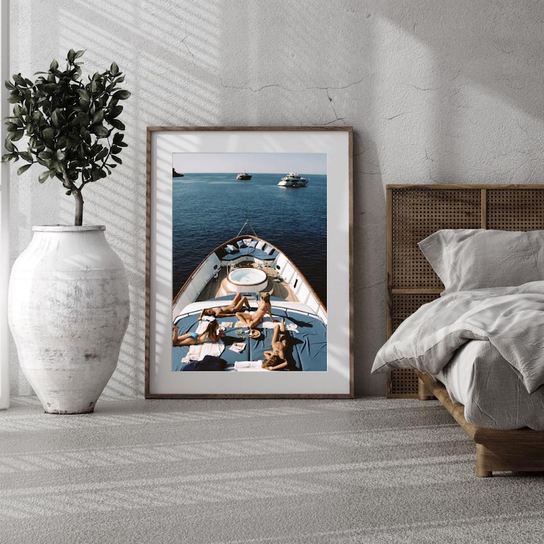 May include: A framed print depicts a yacht on the ocean with people sunbathing on the deck. The yacht has a blue deck with a white jacuzzi. The ocean is a deep blue, and other boats are visible in the distance.