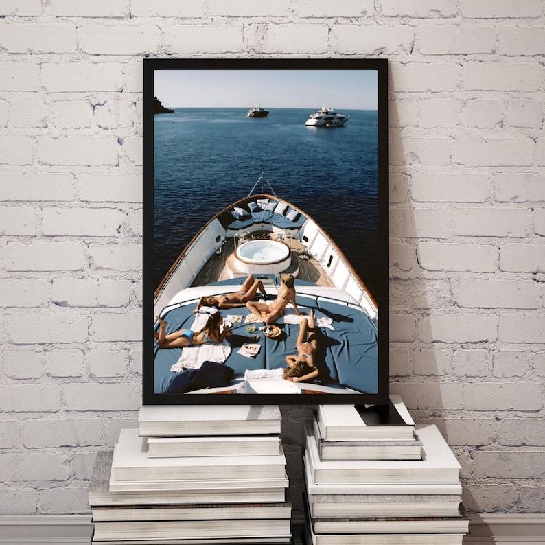 May include: A framed print depicting a yacht with people sunbathing on the deck. The yacht is on the water with other boats in the distance. The deck has a blue cover and a small jacuzzi. The print is displayed on a stack of books.