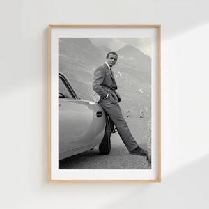 May include: A black and white photographic print featuring a man in a suit leaning against the side of a car. The man is positioned in profile, looking toward the camera, with a mountainous landscape visible in the background behind the vehicle.