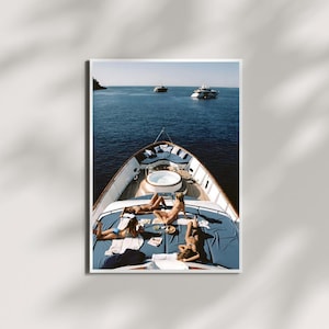 May include: A photograph of a yacht deck with four people sunbathing on blue cushions. The deck features a small jacuzzi and is surrounded by the blue ocean. Two other yachts are visible in the distance.