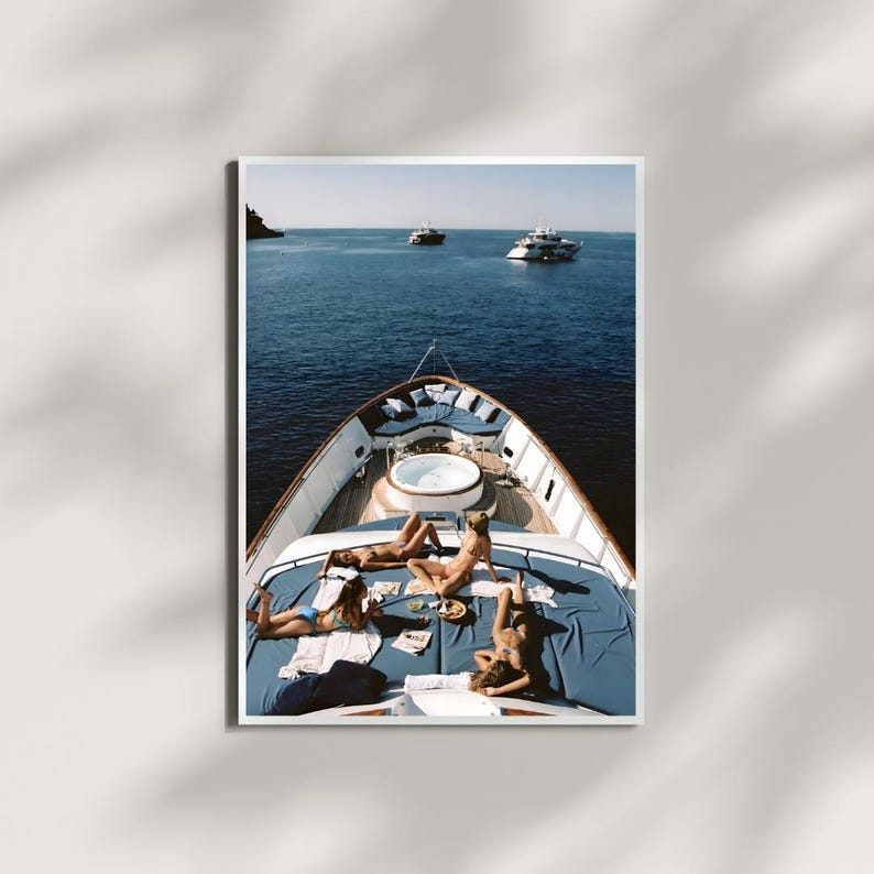 May include: A photograph of a yacht deck with four people sunbathing on blue cushions. The deck features a small jacuzzi and is surrounded by the blue ocean. Two other yachts are visible in the distance.