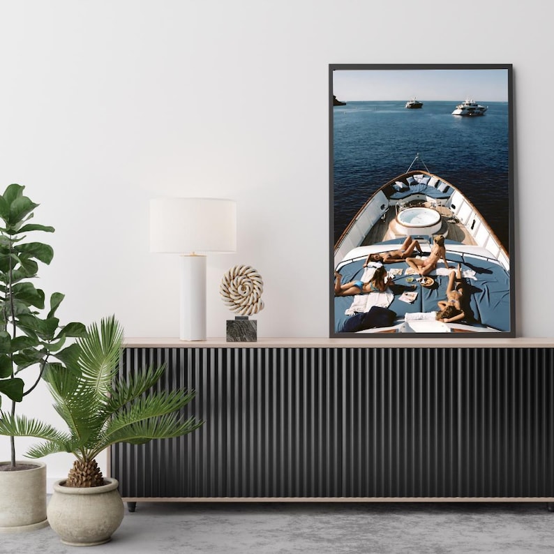 May include: A framed print depicts a yacht's deck with people sunbathing on blue cushions. The ocean and other boats are visible in the background. The artwork is displayed above a black cabinet with a white lamp and decorative objects.