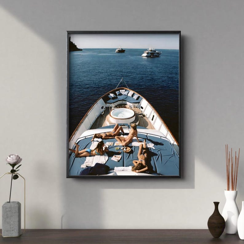 May include: A framed print depicts a yacht with people sunbathing on the deck. The yacht is on the water with other boats in the distance. The deck has a blue cover and a white jacuzzi.