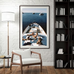 May include: Framed print of a yacht at sea. The yacht's deck features people sunbathing on blue cushions. The ocean is a deep blue, and two other yachts are visible in the distance. The image is displayed on a white brick wall.
