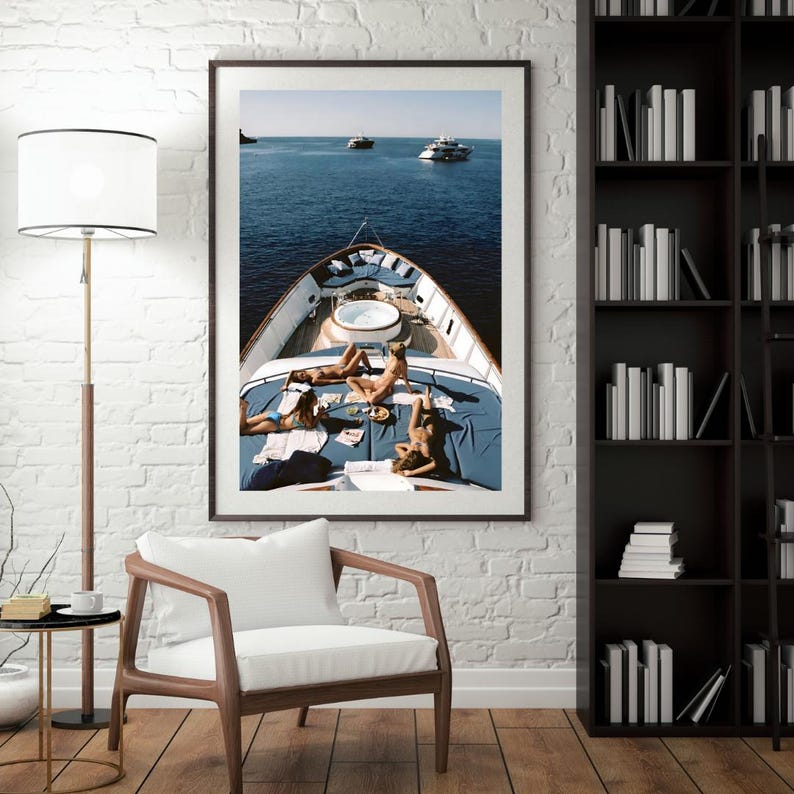 May include: Framed print of a yacht at sea. The yacht's deck features people sunbathing on blue cushions. The ocean is a deep blue, and two other yachts are visible in the distance. The image is displayed on a white brick wall.