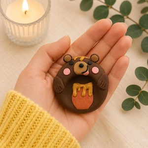 May include: A brown bear-shaped clay figure with a honey pot design. The bear has pink cheeks, a black nose, and a yellow honey accent. The figure is held in a hand, with a lit candle and greenery in the background.