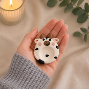 May include: A white and black cow-shaped trinket dish with brown horns and black spots. The dish is held in a hand, with a lit candle in a glass holder and green leaves in the background.