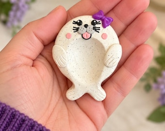 Könnte beinhalten: Eine kleine, weiße, gesprenkelte robbenförmige Ablageschale mit einer glatten, konkaven Mitte. Die Robbe hat eine violette Schleife auf dem Kopf, schwarze Augen, eine schwarze Nase, Schnurrhaare und eine kleine herausgestreckte rosa Zunge.