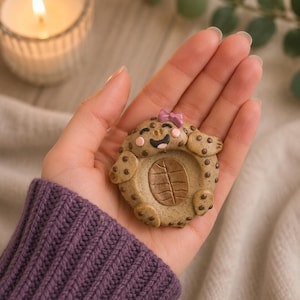 Peut inclure: Figurine de tortue en argile beige faite à la main, avec des taches marron et un nœud rose. La tortue a un visage souriant et un motif en forme de feuille sur sa carapace. La figurine est tenue dans une main, avec une bougie allumée et de la verdure en arrière-plan.