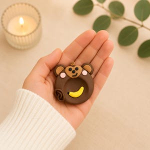 May include: A brown monkey-shaped clay dish with a yellow banana and heart-shaped face is held in a hand. The monkey has tan ears and pink cheeks. The dish is round with a small opening. A lit candle in a glass holder is in the background.