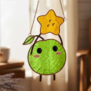 May include: A stained glass suncatcher depicting a green apple character holding a yellow star. The apple has black eyes and pink cheeks. The suncatcher hangs from a chain and is displayed in front of a window.