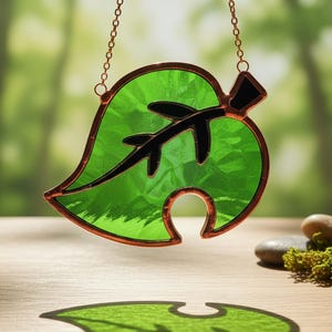 May include: A stained glass leaf in shades of green and black, with a copper-colored border, hangs from a chain. The leaf's design casts a shadow on the wooden surface below. The background is blurred, suggesting a natural outdoor setting.