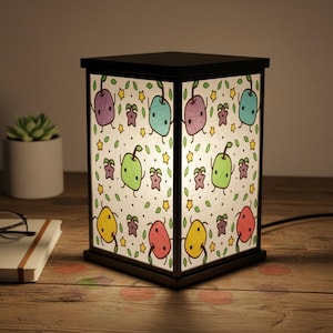 Könnte beinhalten: Eine schwarze, würfelförmige Lampe mit einem skurrilen Design. Die Paneele der Lampe zeigen ein sich wiederholendes Muster aus bunten, cartoonartigen Früchten und Sternen auf weißem Hintergrund. Die Lampe steht auf einer Holzoberfläche.