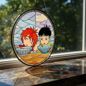 May include: Circular stained glass artwork depicting two anime-style figures enjoying a meal. The design incorporates vibrant colours, including blue, red, yellow, and purple. The glass is framed with a black border and a gold chain for hanging, suitable for a window.