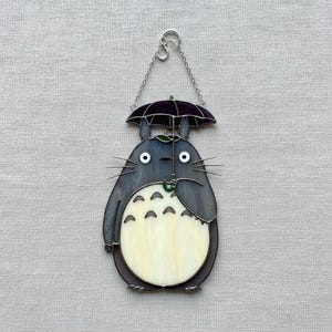 Peut inclure: D&eacute;coration en vitrail repr&eacute;sentant un personnage de dessin anim&eacute; avec un corps gris fonc&eacute;, tenant un parapluie vert. Le personnage a de grands yeux blancs et un ventre cr&egrave;me. Une cha&icirc;ne argent&eacute;e est attach&eacute;e pour l'accrocher.
