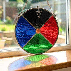 Pode incluir: Um apanhador de sol de vitral circular, dividido em quatro secções: preto, azul, vermelho e verde. Um círculo cor de cobre está no centro e uma seta prateada no topo. O apanhador de sol é pendurado por uma corrente.