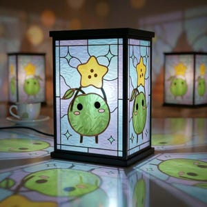 Könnte beinhalten: Eine schwarz gerahmte Buntglaslampe mit einem Cartoon-Apfel-Charakter mit einem gelben Stern. Die Lampe hat eine rechteckige Form und ist beleuchtet, mit zwei weiteren Lampen im Hintergrund. Der Apfel-Charakter ist grün mit schwarzen Augen und einem lächelnden Mund.