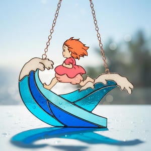 以下が含まれることがあります： 赤い髪とピンクのドレスを着た漫画の女の子が、青と白の波の上を走っているステンドグラスのサンキャッチャー。サンキャッチャーは銅のチェーンで吊り下げられており、背景は青い空です。