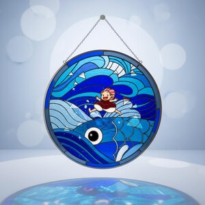 May include: Circular stained glass window art depicting a cartoon girl riding a blue fish through ocean waves. The artwork uses shades of blue and white, with a chain for hanging. The girl has pink hair and a red dress.