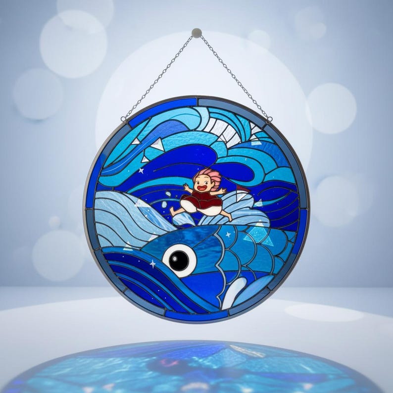May include: Circular stained glass window art depicting a cartoon girl riding a blue fish through ocean waves. The artwork uses shades of blue and white, with a chain for hanging. The girl has pink hair and a red dress.