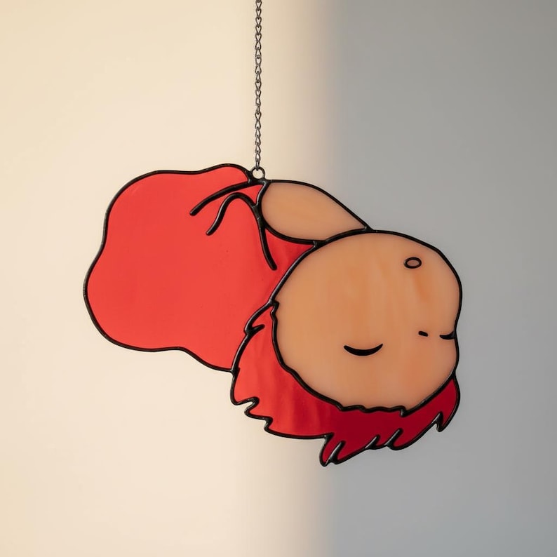 May include: Stained glass artwork of a character with red hair and a red garment, suspended by a chain. The character's face is light orange with closed eyes. Black lines outline the shapes and details of the artwork.
