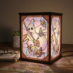 May include: A stained glass lamp with a wooden frame. The lamp features a whimsical design of a white creature with a long beak, surrounded by small figures in wizard hats. The glass has a soft pink and purple gradient.