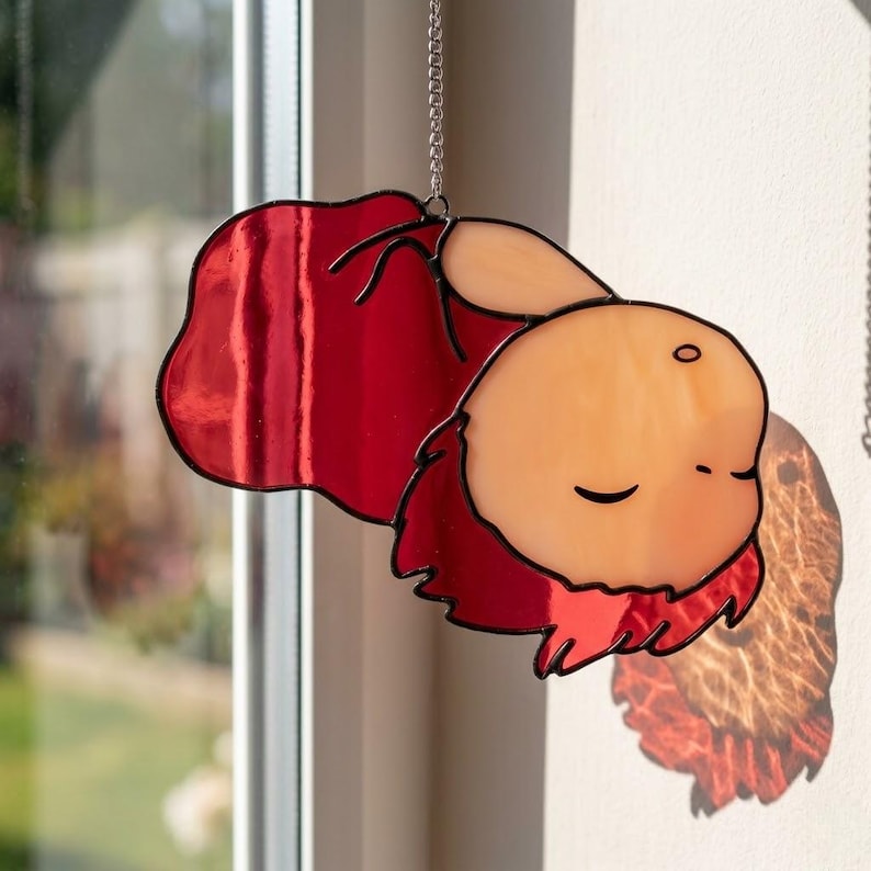 May include: A stained glass suncatcher featuring a cartoon character with closed eyes and a red cape. The character's face is peach-coloured with black outlines, and the cape is a vibrant red. A silver chain is attached for hanging.