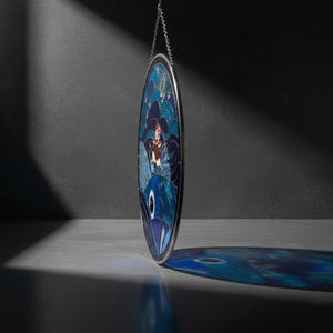 May include: An oval stained glass panel depicting a nautical scene. It features a woman with pink hair and a blue fish, set against blue waves. The panel is framed in silver and suspended by a chain.