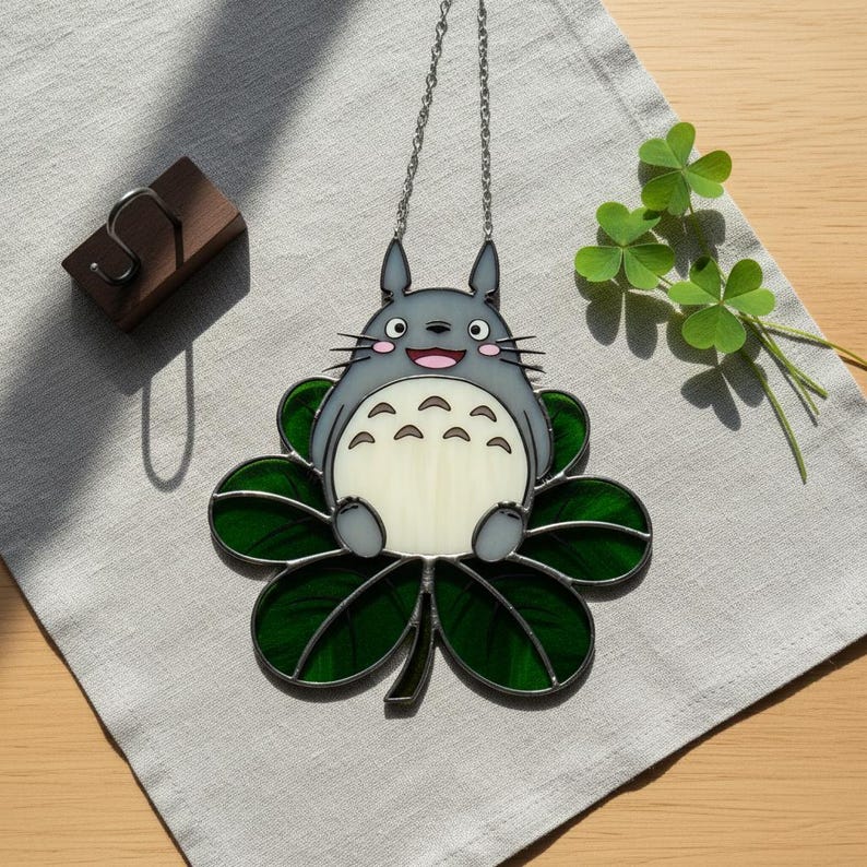 Peut inclure: Suncatcher en vitrail repr&eacute;sentant un personnage Totoro gris assis sur un tr&egrave;fle &agrave; quatre feuilles vert. Il est suspendu &agrave; une cha&icirc;ne argent&eacute;e. Le design comprend un Totoro souriant avec un ventre blanc et un tr&egrave;fle vert.