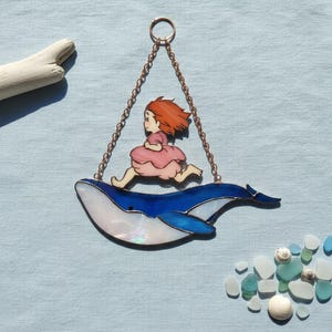 May include: Stained glass artwork showing a girl running on a blue whale. The whale has a white and blue iridescent finish. The girl wears a pink dress and has red hair. The piece is suspended by a copper chain, ideal for a window.