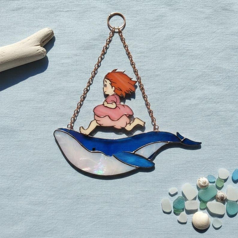 May include: Stained glass artwork showing a girl running on a blue whale. The whale has a white and blue iridescent finish. The girl wears a pink dress and has red hair. The piece is suspended by a copper chain, ideal for a window.