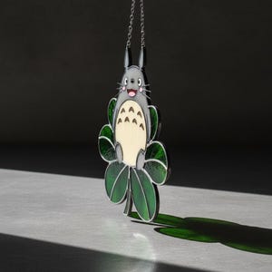Peut inclure: Pendentif Totoro gris et cr&egrave;me, orn&eacute; de feuilles vertes. Le pendentif est suspendu &agrave; une cha&icirc;ne argent&eacute;e. Le personnage de Totoro souriant est entour&eacute; de feuilles vertes.