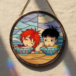 May include: Circular stained glass artwork depicting two anime-style figures eating from bowls. The piece has a brown border and a chain for hanging. The background shows a sky and ocean scene.
