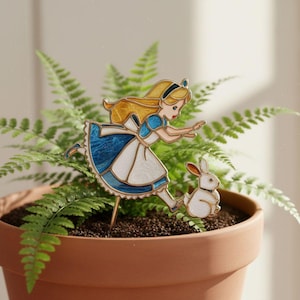 May include: A decorative metal plant stake featuring a colourful enamel illustration of a girl with long blonde hair wearing a blue dress and white apron, reaching towards a small white rabbit with brown ears, mounted on a thin gold-toned metal rod.