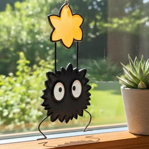 Peut inclure: Ornement d&eacute;coratif suspendu repr&eacute;sentant un personnage noir et &eacute;pineux avec de grands yeux blancs, suspendu sous une &eacute;toile jaune. L'ornement est suspendu devant un arri&egrave;re-plan vert flou. Le design est fantaisiste.