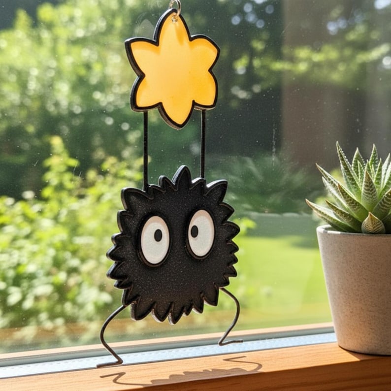 Peut inclure: Ornement d&eacute;coratif suspendu repr&eacute;sentant un personnage noir et &eacute;pineux avec de grands yeux blancs, suspendu sous une &eacute;toile jaune. L'ornement est suspendu devant un arri&egrave;re-plan vert flou. Le design est fantaisiste.