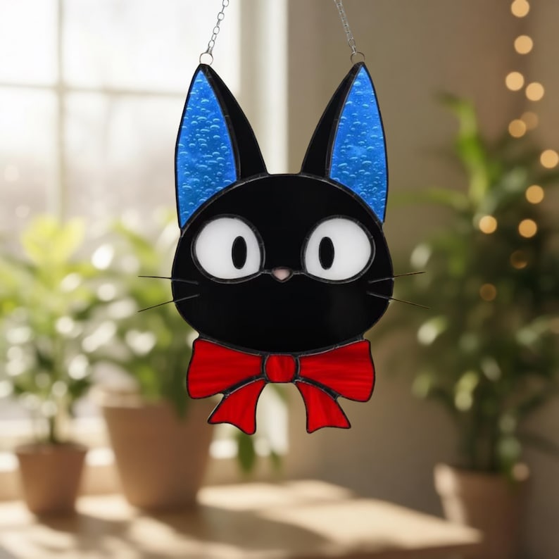 May include: Black cat stained glass art with blue ears and a red bow tie. The cat has large white eyes and whiskers. The cat is hanging from a chain. The background is blurred with plants and a window.