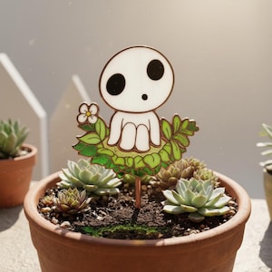 Puede incluir: Una estaca para plantas blanca y verde con un personaje de dibujos animados con grandes ojos negros y una expresión de sorpresa. La estaca está colocada en una maceta de terracota con suculentas. El personaje está rodeado de hojas verdes y una pequeña flor blanca.