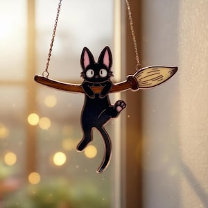 May include: A black cat with pink-lined ears and paws hangs from a brown broom. The cat is suspended by a chain, and the broom has a light-colored brush. The background is blurred with warm tones.