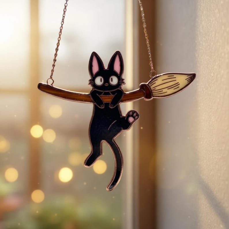 May include: A black cat with pink-lined ears and paws hangs from a brown broom. The cat is suspended by a chain, and the broom has a light-colored brush. The background is blurred with warm tones.