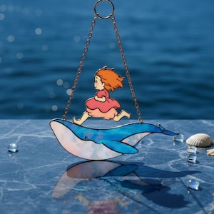 May include: A decorative suncatcher depicting a girl in a pink dress running on a blue whale, suspended by a gold chain. The whale has a white belly. The background is a blurred blue, evoking water. A charming, whimsical design.