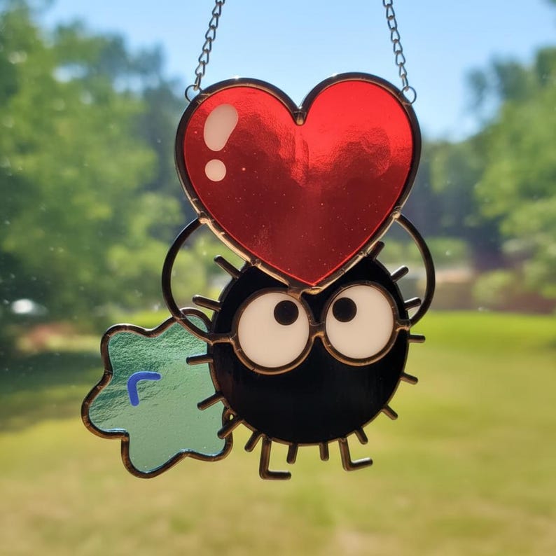 Puede incluir: Atrapasoles de vidriera con un personaje de duende de holl&iacute;n negro con grandes ojos blancos y un coraz&oacute;n rojo. Un elemento verde en forma de estrella est&aacute; a la izquierda. El atrapasoles cuelga de una cadena plateada, ideal para decoraci&oacute;n.