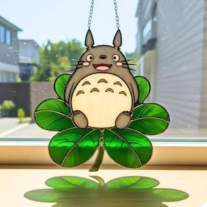 Peut inclure: D&eacute;coration en vitrail repr&eacute;sentant un personnage Totoro gris souriant, assis sur un tr&egrave;fle &agrave; quatre feuilles vert. Le tr&egrave;fle a un aspect textur&eacute;. La d&eacute;coration est suspendue par une cha&icirc;ne et est expos&eacute;e devant une fen&ecirc;tre.