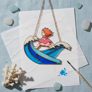 May include: Stained glass artwork showing a girl running on a blue and white wave. It has a copper chain for hanging. The girl wears a pink dress and has red hair. Decorative sea glass and coral are in the background. The artwork is approximately 15 cm in length.