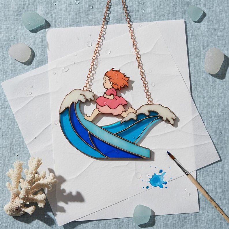 May include: Stained glass artwork showing a girl running on a blue and white wave. It has a copper chain for hanging. The girl wears a pink dress and has red hair. Decorative sea glass and coral are in the background. The artwork is approximately 15 cm in length.